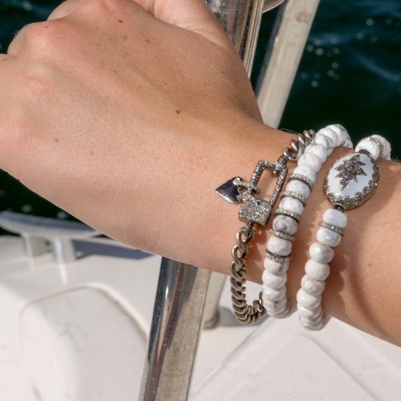May include: Three white beaded bracelets with silver charms, including a heart and a padlock, on a person's wrist.