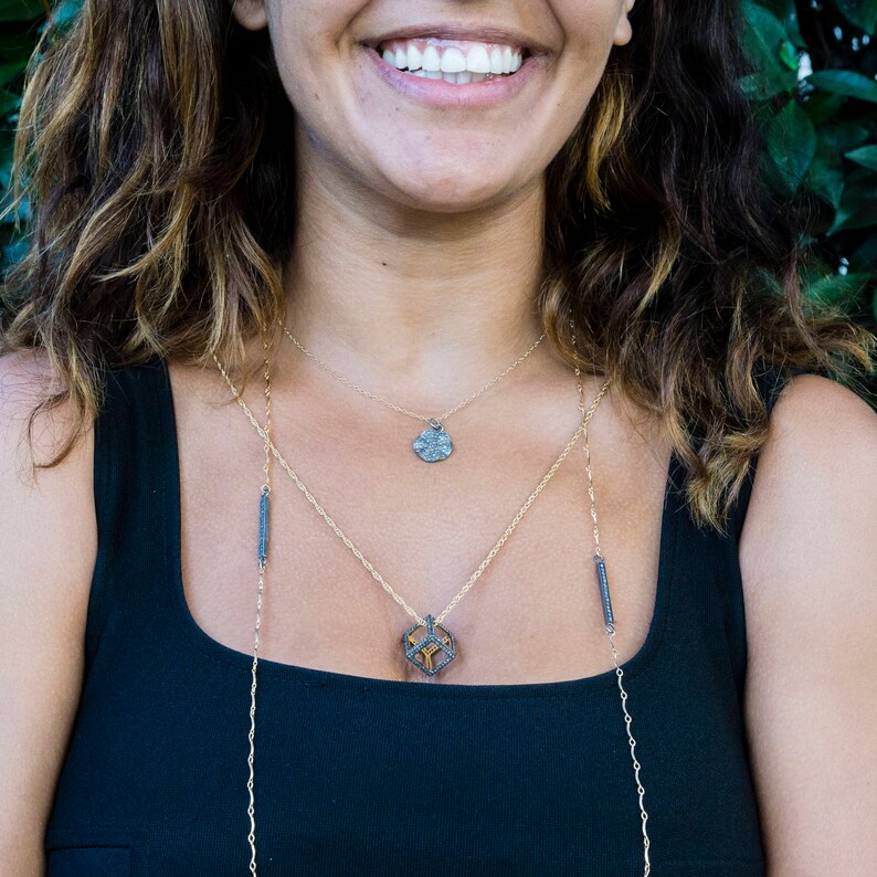 May include: A woman wearing a black tank top with three gold necklaces. The necklaces feature a small round pendant, a geometric pendant, and two thin black bars.