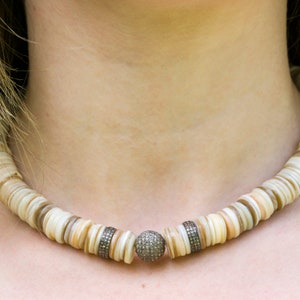 May include: A white and brown shell necklace with a silver and diamond accent. The necklace is made of round, flat shells and has a single silver bead with small diamonds set into it.