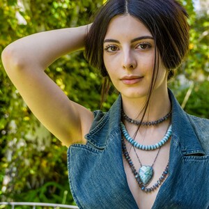 Puede incluir: Una mujer con el pelo largo y oscuro lleva un chaleco vaquero con varias capas de collares que presentan un gran colgante en forma de coraz&oacute;n. Los collares est&aacute;n hechos de cuentas azules, grises y plateadas.