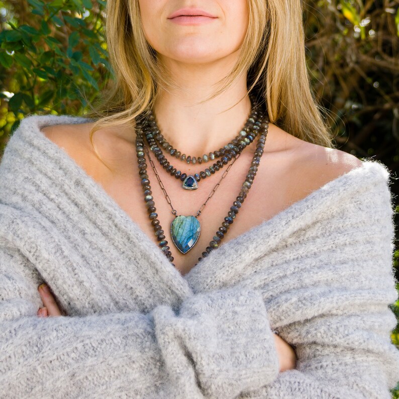 Puede incluir: Una mujer lleva un su&eacute;ter de punto gris con un collar de capas que presenta un colgante en forma de coraz&oacute;n con una piedra preciosa azul y verde. El collar tambi&eacute;n incluye un colgante m&aacute;s peque&ntilde;o con una piedra preciosa azul.