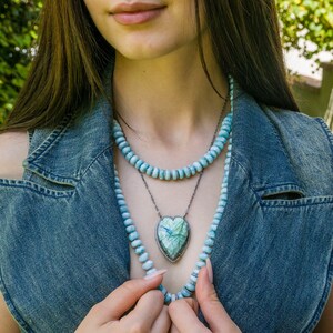 Puede incluir: Una mujer lleva un chaleco vaquero con un collar de capas que presenta un colgante en forma de coraz&oacute;n con una piedra preciosa verde y azul. El collar est&aacute; hecho de cuentas de color azul claro.