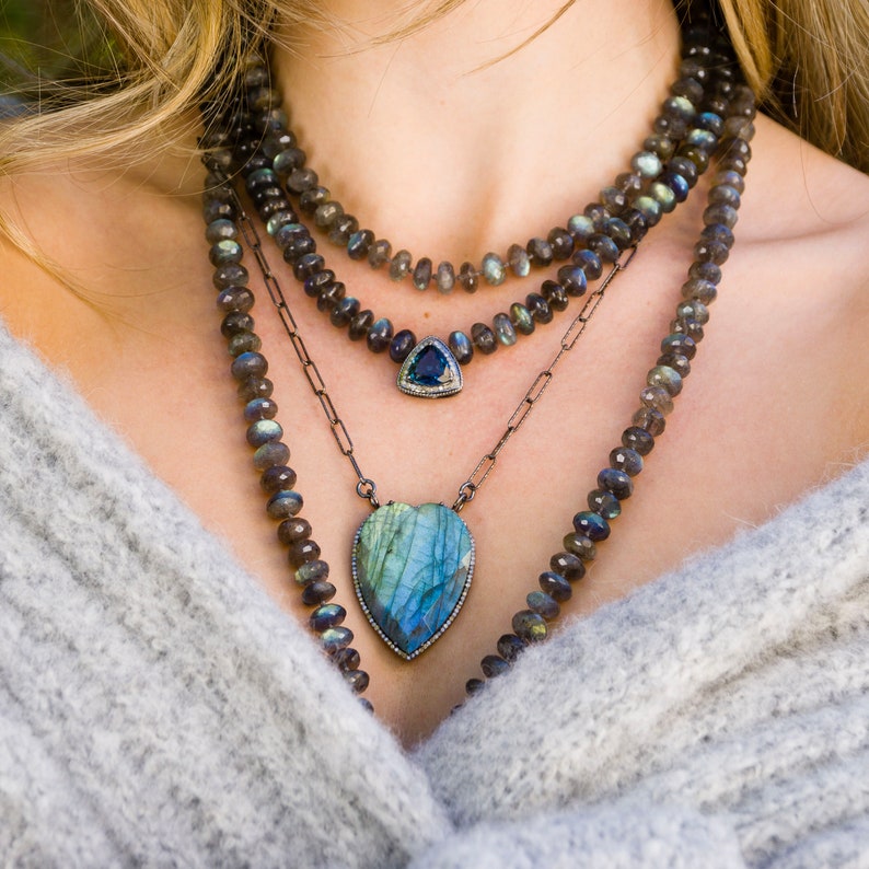 Puede incluir: Un collar de capas con tres colgantes. El colgante superior es una piedra preciosa azul triangular con un engaste de plata. El colgante del medio es una cadena de plata con un peque&ntilde;o dije de plata. El colgante inferior es una piedra preciosa azul en forma de coraz&oacute;n con un engaste de plata.