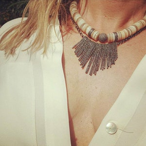 May include: A white and brown shell necklace with a silver pendant featuring a fringe of small silver beads. The necklace is worn by a person with a white shirt.