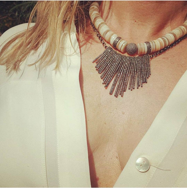 May include: A white and brown shell necklace with a silver pendant featuring a fringe of small silver beads. The necklace is worn by a person with a white shirt.