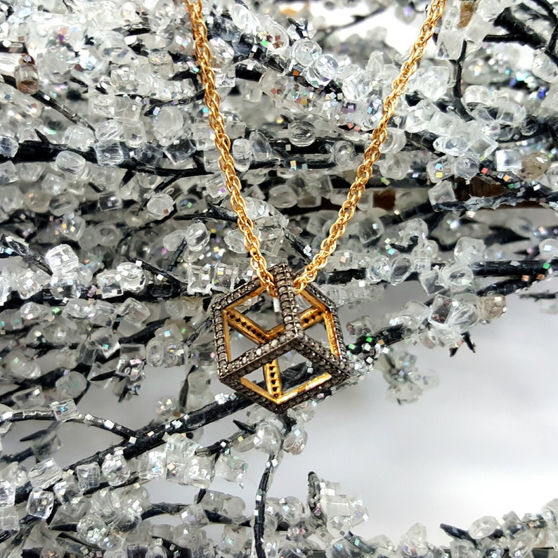 May include: A gold chain necklace with a diamond-encrusted cube pendant. The cube is made of gold and has a geometric design. The necklace is photographed against a sparkly background.