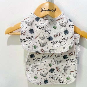 May include: Two white cotton baby bibs with a wizard theme. The bibs have a black and white print of a wizard, a cauldron, a wand, glasses, and stars. The text on the bibs reads "Wizard in Training".