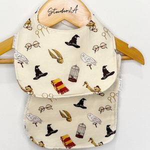 May include: Two white cotton baby bibs with a Harry Potter theme. The bibs feature a white background with a repeating pattern of black hats, owls, glasses, golden snitches, and cages. The bibs are hanging on a wooden hanger.