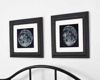 Op de afbeelding: Twee zwart ingelijste prints van een maan in verschillende fasen, hangend aan een witte muur boven een zwart metalen hoofdeinde.