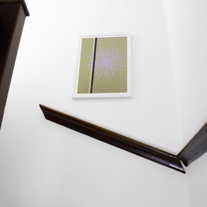 Op de afbeelding: Een ingelijste abstracte print met een witte lijst en een lichtgroene achtergrond. Een paars en wit geometrisch ontwerp bevindt zich in het midden van de print. De print hangt aan een witte muur.