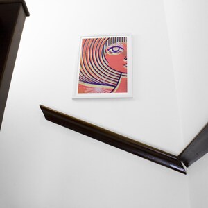 May include: A framed print of a colorful abstract portrait of a woman with a large eye and wavy hair. The background is a light orange color.