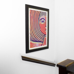May include: A framed abstract art print of a woman's face with colorful stripes and a single eye. The background is a light orange color.