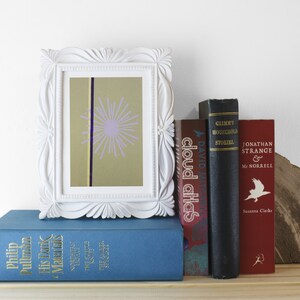 Op de afbeelding: Een wit fotolijstje met een lichtgroen en paars abstract ontwerp erin. Het lijstje staat op een plank met een stapel boeken eronder. De boeken hebben titels als "Cloud Atlas" en "Jonathan Strange & Mr Norrell".