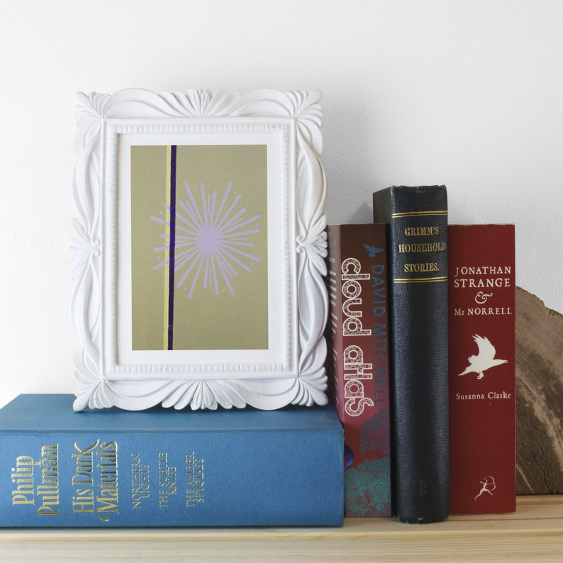 Op de afbeelding: Een wit fotolijstje met een lichtgroen en paars abstract ontwerp erin. Het lijstje staat op een plank met een stapel boeken eronder. De boeken hebben titels als "Cloud Atlas" en "Jonathan Strange & Mr Norrell".