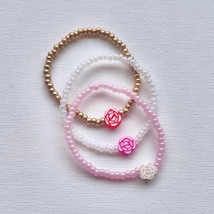 May include: Three beaded bracelets in gold, white, and pink. The pink bracelets feature rose-shaped charms in red, pink, and white. The bracelets are elastic and appear to be made of small beads.