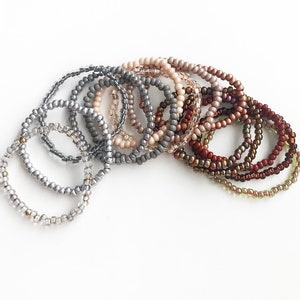 May include: A set of 12 beaded bracelets in various shades of gray, brown, and beige. The bracelets are stacked on top of each other, creating a gradient effect.