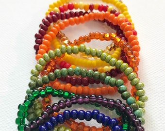 May include: A stack of colorful beaded bracelets in shades of green, orange, red, purple, and blue. The bracelets are made of small, round beads and are arranged in a rainbow pattern.