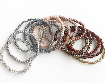 May include: A set of 12 beaded bracelets in various shades of gray, brown, and beige. The bracelets are stacked on top of each other, creating a gradient effect.