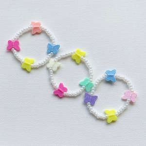 May include: Three white pearl bracelets with colorful butterfly charms. The bracelets are arranged in a circle shape, with the butterflies facing outwards. The butterflies are in various colors, including pink, yellow, blue, green, and purple.