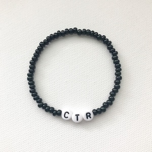 May include: Black beaded bracelet with white letter beads spelling "CTR". The bracelet is made of small, round black beads strung together. The white letter beads are centered on the bracelet.
