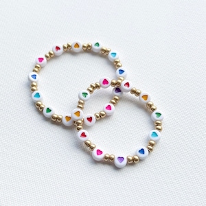 May include: Two bracelets with white beads and rainbow heart charms. The bracelets are made with gold beads and are layered on top of each other.
