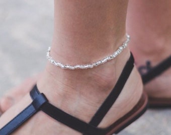 Puede incluir: Un tobillo de plata con un delicado diseño de cadena en el tobillo de una persona. El tobillo se lleva con sandalias negras.