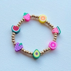 May include: A gold beaded bracelet with colorful fruit charms. The charms include a strawberry, lemon, watermelon, grapes, avocado, and grapefruit.