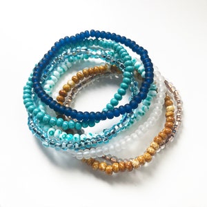 May include: A set of seven beaded bracelets in shades of blue, turquoise, and brown. The bracelets are made of small, round beads and are stacked on top of each other.