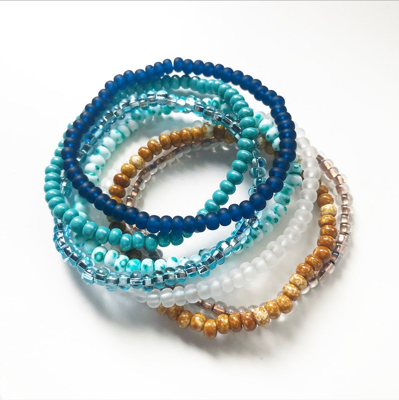 May include: A set of seven beaded bracelets in shades of blue, turquoise, and brown. The bracelets are made of small, round beads and are stacked on top of each other.