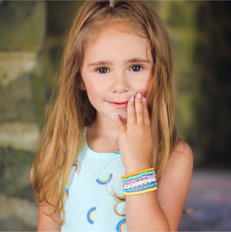 May include: A young girl with long blonde hair is wearing a light blue and white dress with rainbow patterns. She is wearing a stack of colorful beaded bracelets on her wrist.