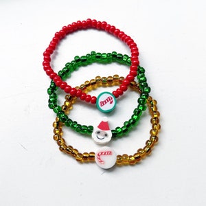 Puede incluir: Tres pulseras de cuentas coloridas con dijes navide&ntilde;os. Las pulseras son rojas, verdes y doradas. Los dijes son un bast&oacute;n de caramelo, un Pap&aacute; Noel y un bast&oacute;n de caramelo.