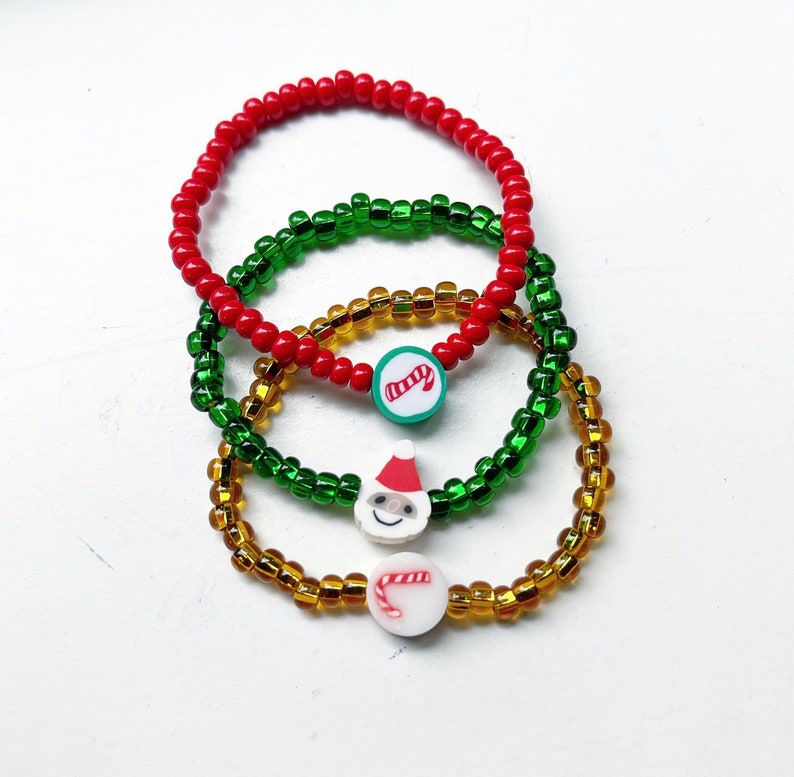 Puede incluir: Tres pulseras de cuentas coloridas con dijes navide&ntilde;os. Las pulseras son rojas, verdes y doradas. Los dijes son un bast&oacute;n de caramelo, un Pap&aacute; Noel y un bast&oacute;n de caramelo.