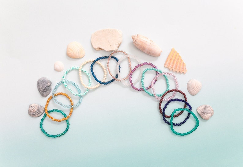 May include: A collection of colorful beaded bracelets arranged in a semi-circle on a light blue background. The bracelets are made of various colors, including blue, green, purple, and brown. There are also several seashells scattered around the bracelets.