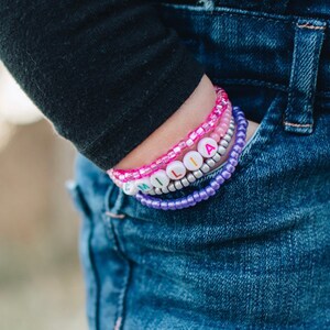 May include: A close-up of a person's wrist wearing a stack of colorful beaded bracelets. The bracelets spell out the name "MILIA" in white letters with rainbow accents.