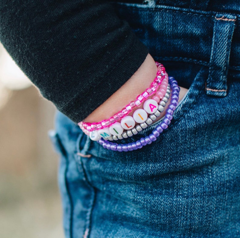 May include: A close-up of a person's wrist wearing a stack of colorful beaded bracelets. The bracelets spell out the name "MILIA" in white letters with rainbow accents.