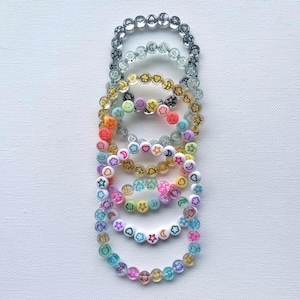 May include: A stack of colorful beaded bracelets with different charms, including stars, hearts, clouds, and moons. The bracelets are made of clear glass beads with a rainbow of colors.