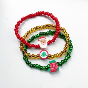 Puede incluir: Tres pulseras con tem&aacute;tica navide&ntilde;a hechas con cuentas rojas, verdes y doradas. Cada pulsera tiene un dije diferente: Santa Claus, un &aacute;rbol de Navidad y una media.