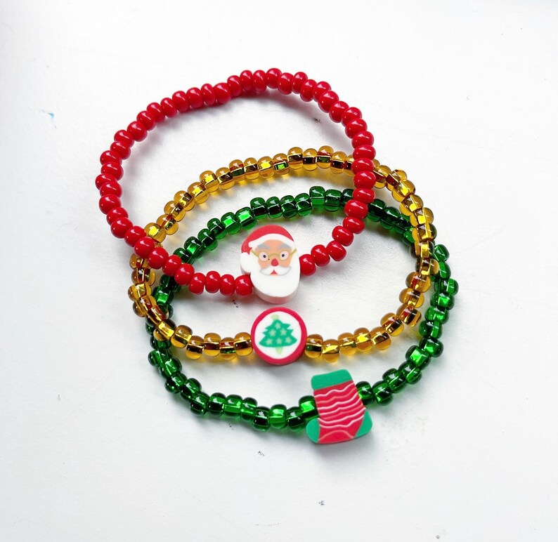 Puede incluir: Tres pulseras con tem&aacute;tica navide&ntilde;a hechas con cuentas rojas, verdes y doradas. Cada pulsera tiene un dije diferente: Santa Claus, un &aacute;rbol de Navidad y una media.