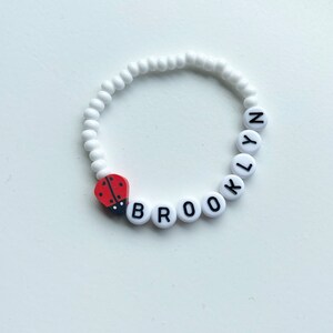K&ouml;nnte beinhalten: Wei&szlig;es Perlenarmband mit dem Namen "BROOKLYN" in schwarzen Buchstaben. Ein roter Marienk&auml;fer-Anh&auml;nger ist am Armband befestigt.