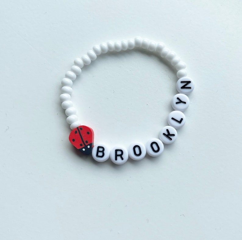 K&ouml;nnte beinhalten: Wei&szlig;es Perlenarmband mit dem Namen "BROOKLYN" in schwarzen Buchstaben. Ein roter Marienk&auml;fer-Anh&auml;nger ist am Armband befestigt.