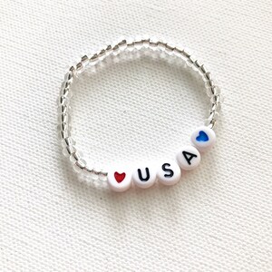 May include: A white beaded bracelet with the letters "USA" and a red heart bead. A blue heart bead is on the other side of the bracelet.