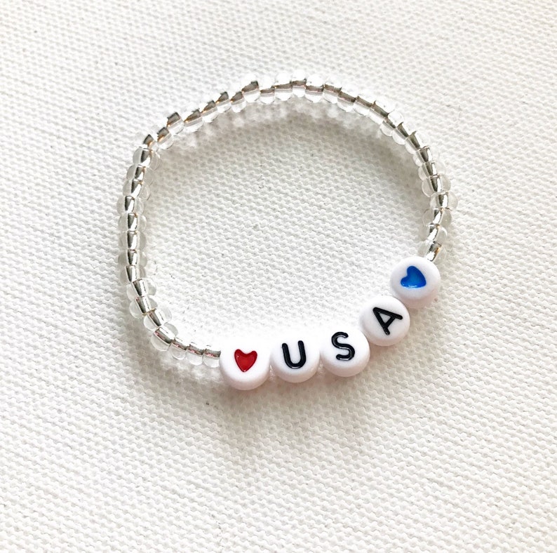 May include: A white beaded bracelet with the letters "USA" and a red heart bead. A blue heart bead is on the other side of the bracelet.