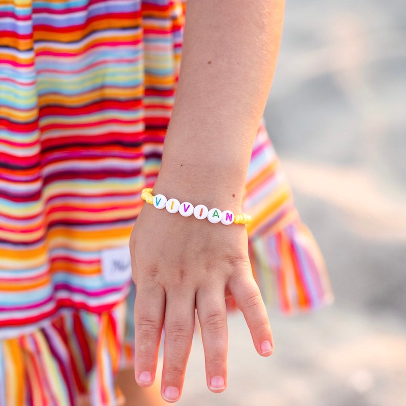 May include: A yellow beaded bracelet with white letter beads spelling out the name "VIVIAN".