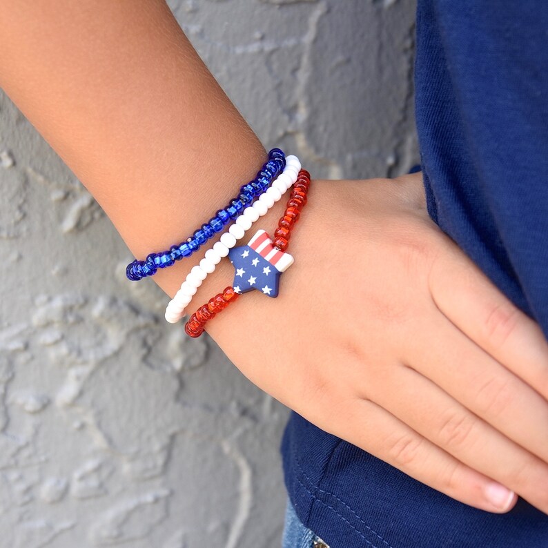 May include: A red, white, and blue beaded bracelet with a star-shaped charm. The bracelet is made of small, round beads and is worn on a person's wrist.