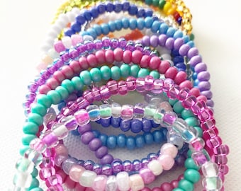 May include: A stack of colorful beaded bracelets in shades of pink, purple, blue, green, and white. The bracelets are made of small, round beads and are arranged in a rainbow pattern.