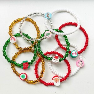 Puede incluir: Un conjunto de pulseras de cuentas coloridas con dijes de tem&aacute;tica navide&ntilde;a. Los dijes incluyen un mu&ntilde;eco de nieve, una media, un bast&oacute;n de caramelo, un &aacute;rbol de Navidad y Santa Claus.