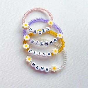 Könnte beinhalten: Vier Armbänder aus Perlen in Rosa, Lila, Gold und Transparent. Jedes Armband hat weiße Buchstabenperlen mit den Namen: "HAZEL", "VIENNA", "ELISA" und "MAMA". Weiße, gänseblümchenförmige Perlen akzentuieren die Namen.
