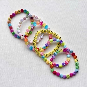 May include: Five colorful beaded bracelets with smiley face beads in various colors, including yellow, pink, purple, and green. The bracelets are arranged on a white surface.