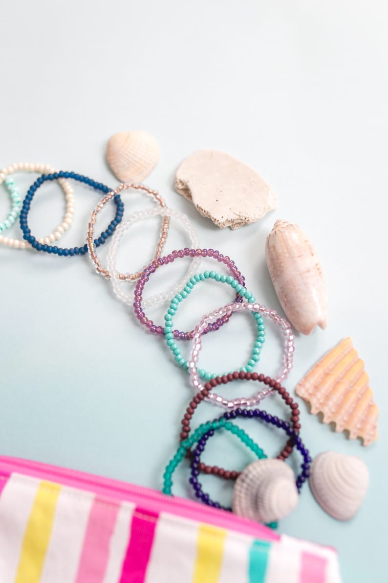 May include: A collection of colorful beaded bracelets with a striped pink and white zippered pouch and seashells on a light blue background.