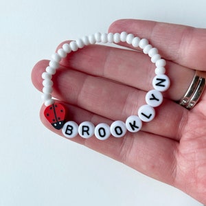K&ouml;nnte beinhalten: Wei&szlig;es Perlenarmband mit dem Namen "Brooklyn", der in schwarzen Buchstaben auf jeder Perle geschrieben ist. Ein roter und schwarzer Marienk&auml;fer-Anh&auml;nger ist am Armband befestigt.